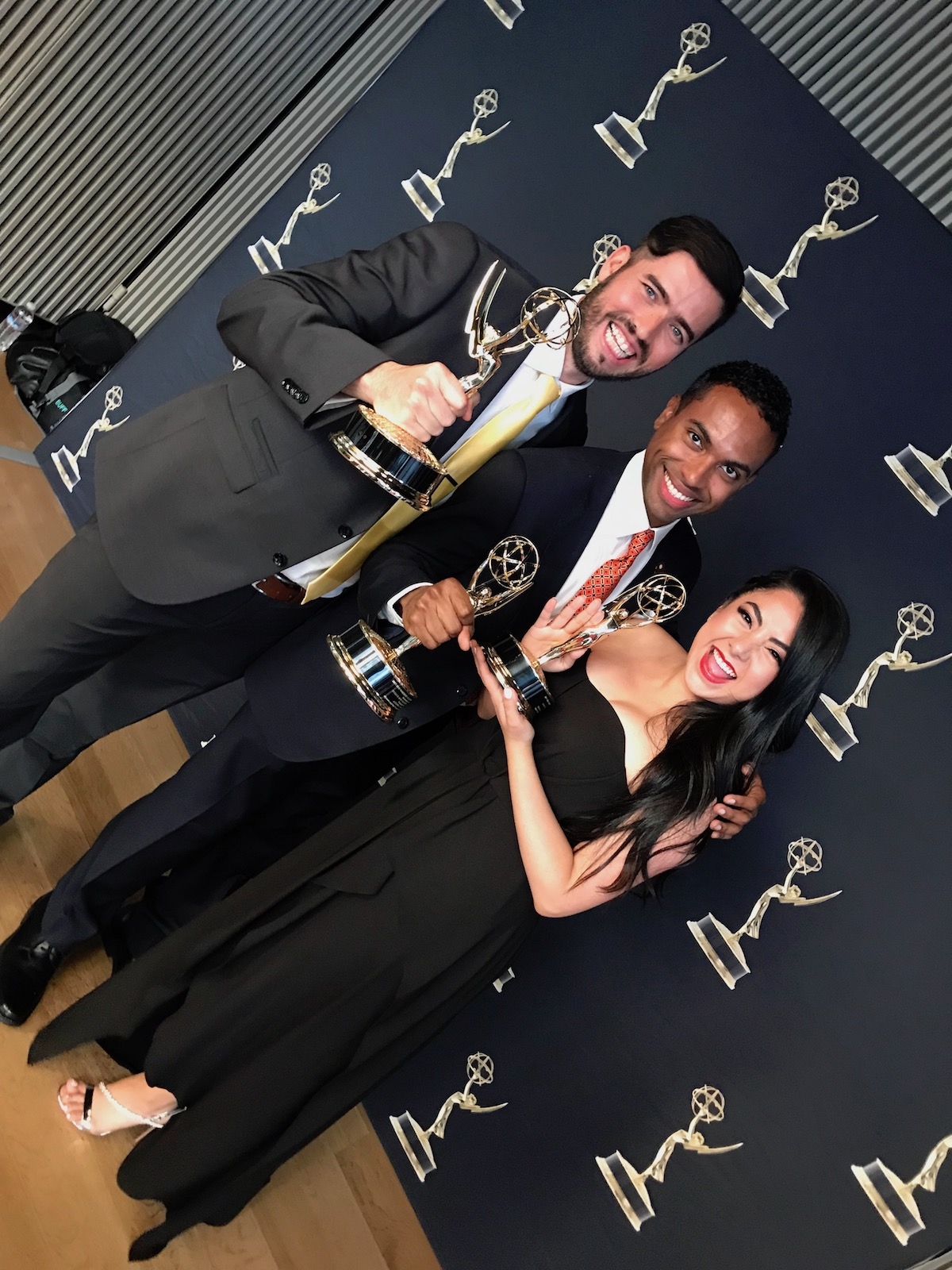 Antonio Ayala (center) holds EMMY trophies with one person on each side in front of EMMY step and repeat